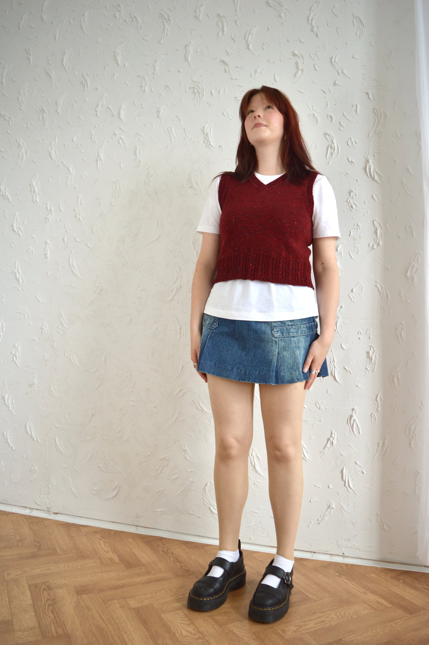 Full-length image of model wearing The Cable Vest in red, styled with a white T-shirt, denim mini skirt, and black shoes.