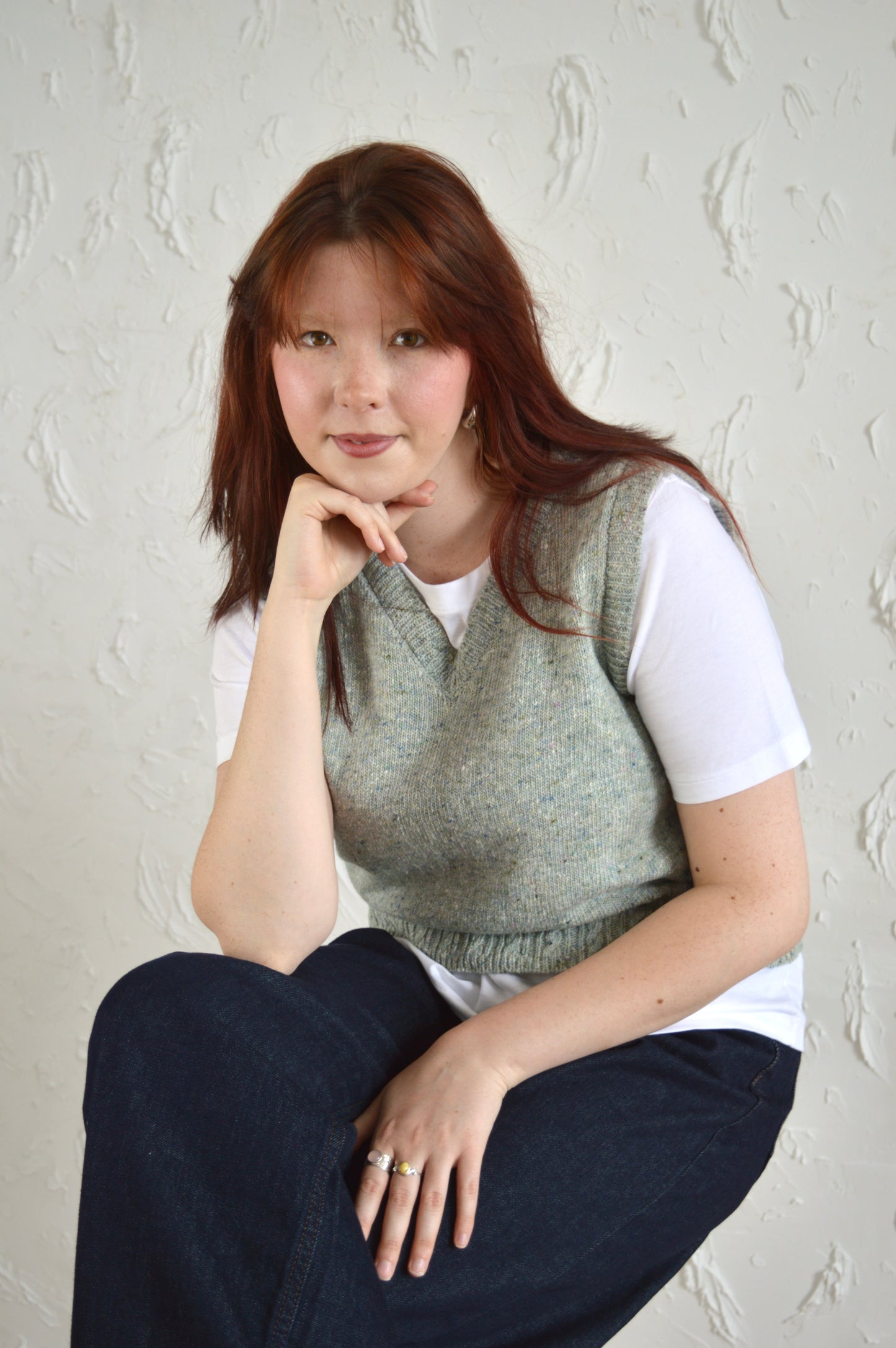 Model wearing The Cable Vest, seated on a stool with hand resting on chin in a relaxed pose.