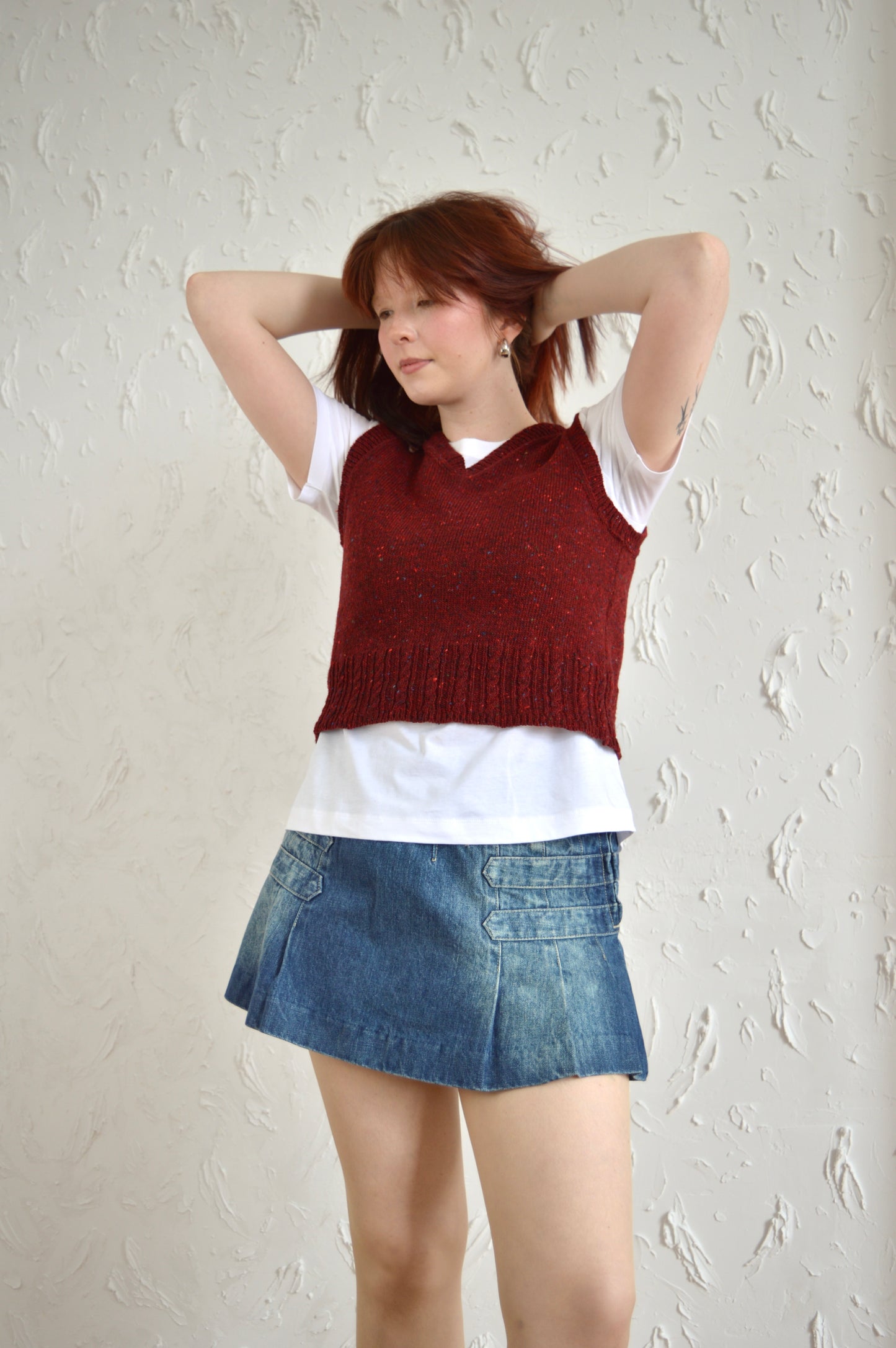 Model in red Cable Vest, posing with hands in her hair.