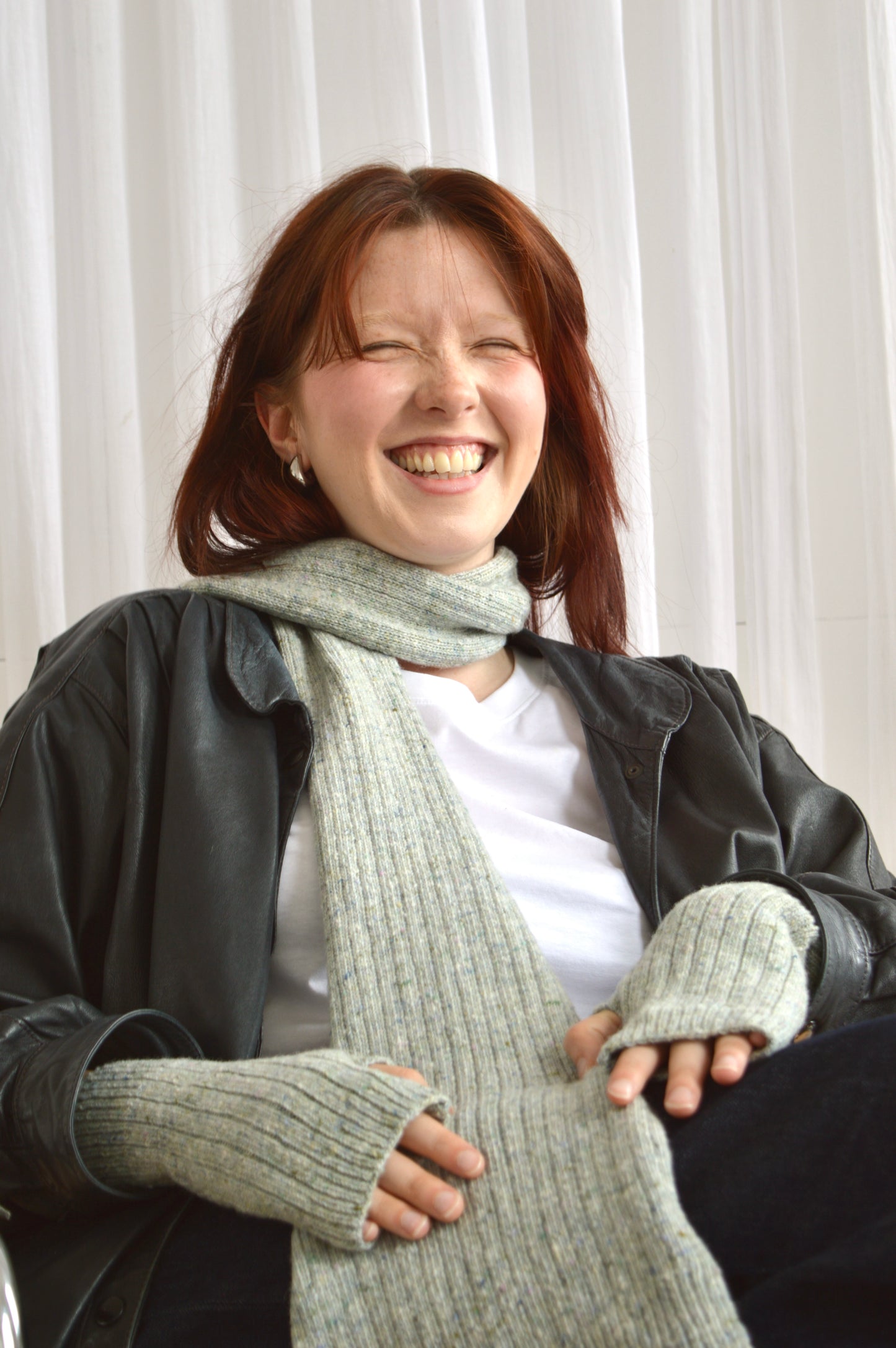 Model wears The Reversible Rib Scarf in light grey, seated and laughing