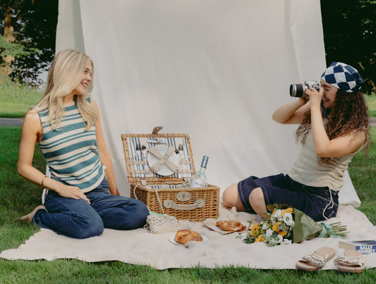 Two women sit at a picnic both wearing The Blanket Top. One woman is taking a picture of the other
