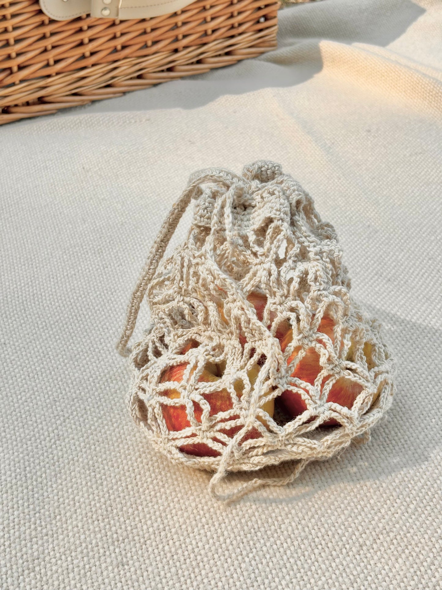 A cream-colored crochet drawstring bag filled with red apples, resting on a textured beige linen blanket with a wicker basket in the background.