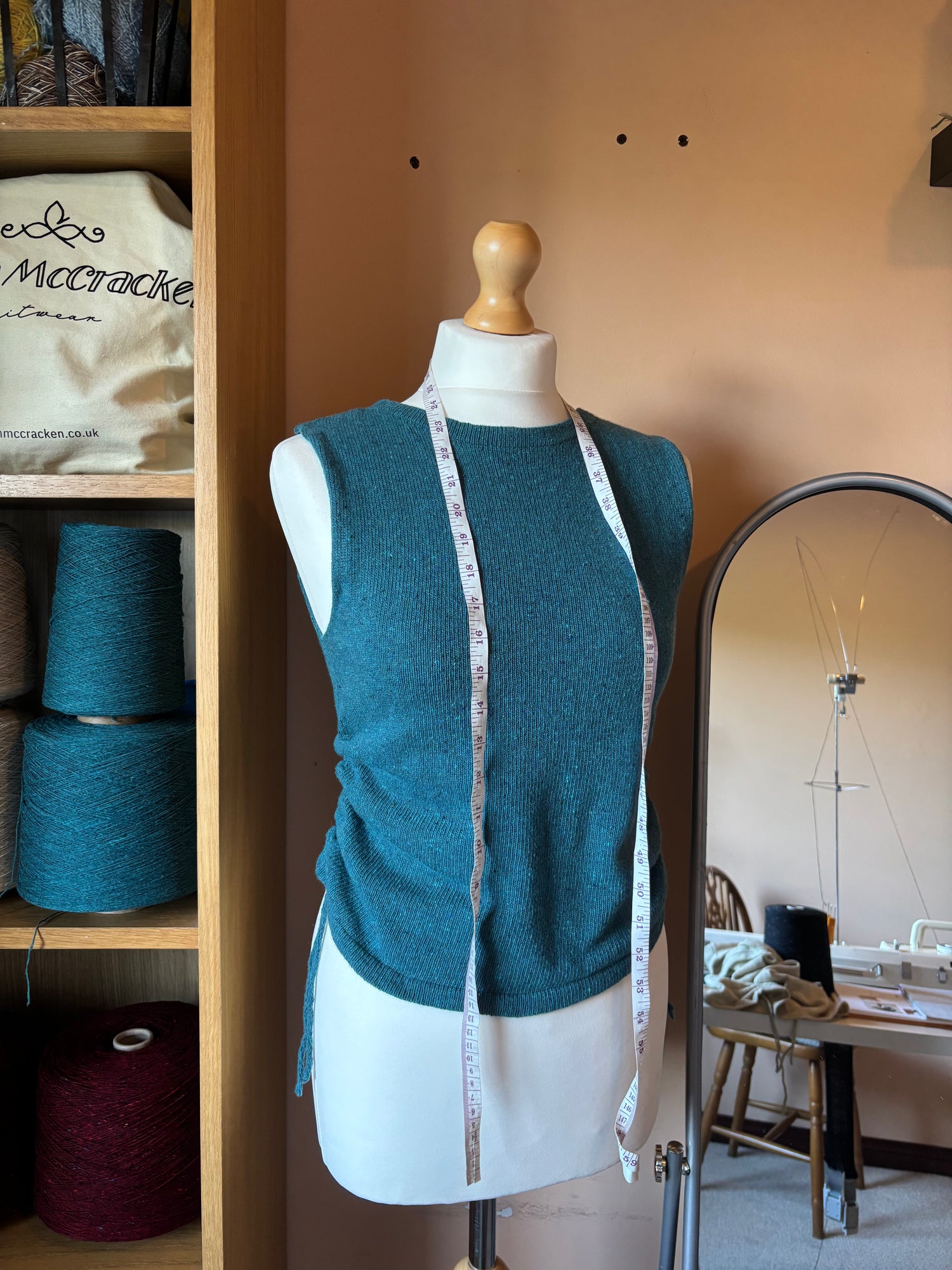 The Blanket Top in teal displayed on a white mannequin torso with a measuring tape draped down the front. The mannequin is set in a knitting studio with shelves of yarn on the left and a mirror on the right.