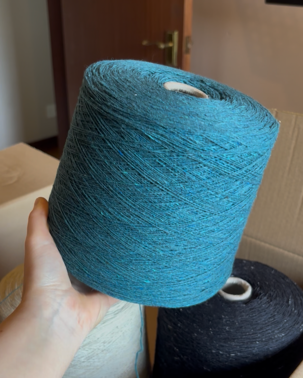 A hand holding a large spool of textured blue yarn, with other yarn spools visible in cardboard boxes in the background.