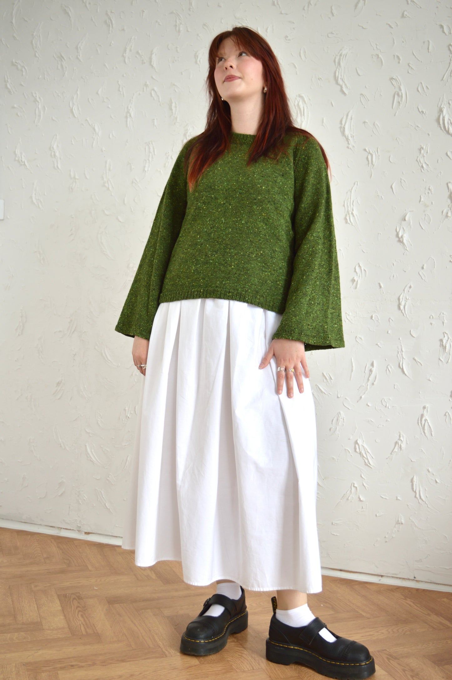 Full length shot of model wearing The Bell Sweater in green, wearing a white skirt and black shoes.