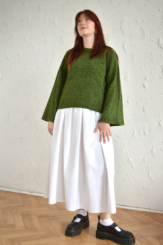 Full length shot of model wearing The Bell Sweater in green, wearing a white skirt and black shoes.