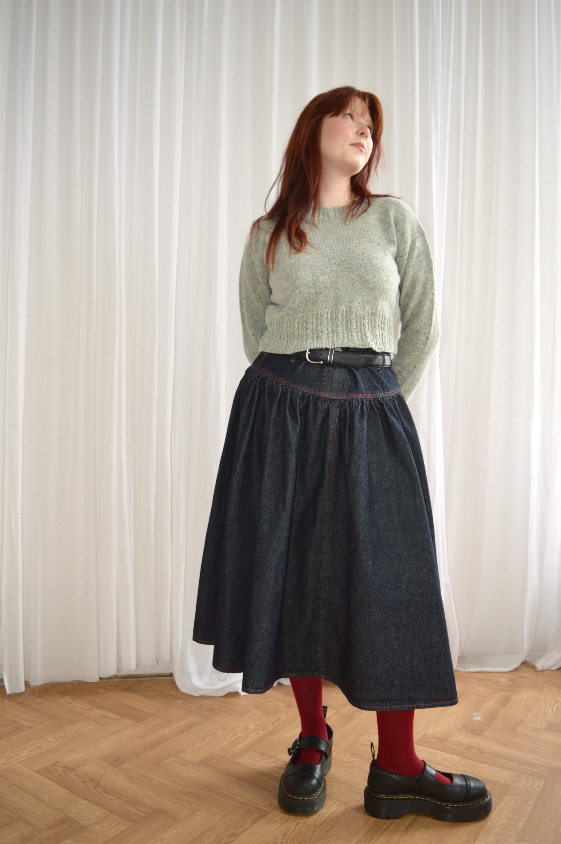Full length view of model wearing the Cable Sweater in light grey and styled with denim skirt, red tights and black shoes.