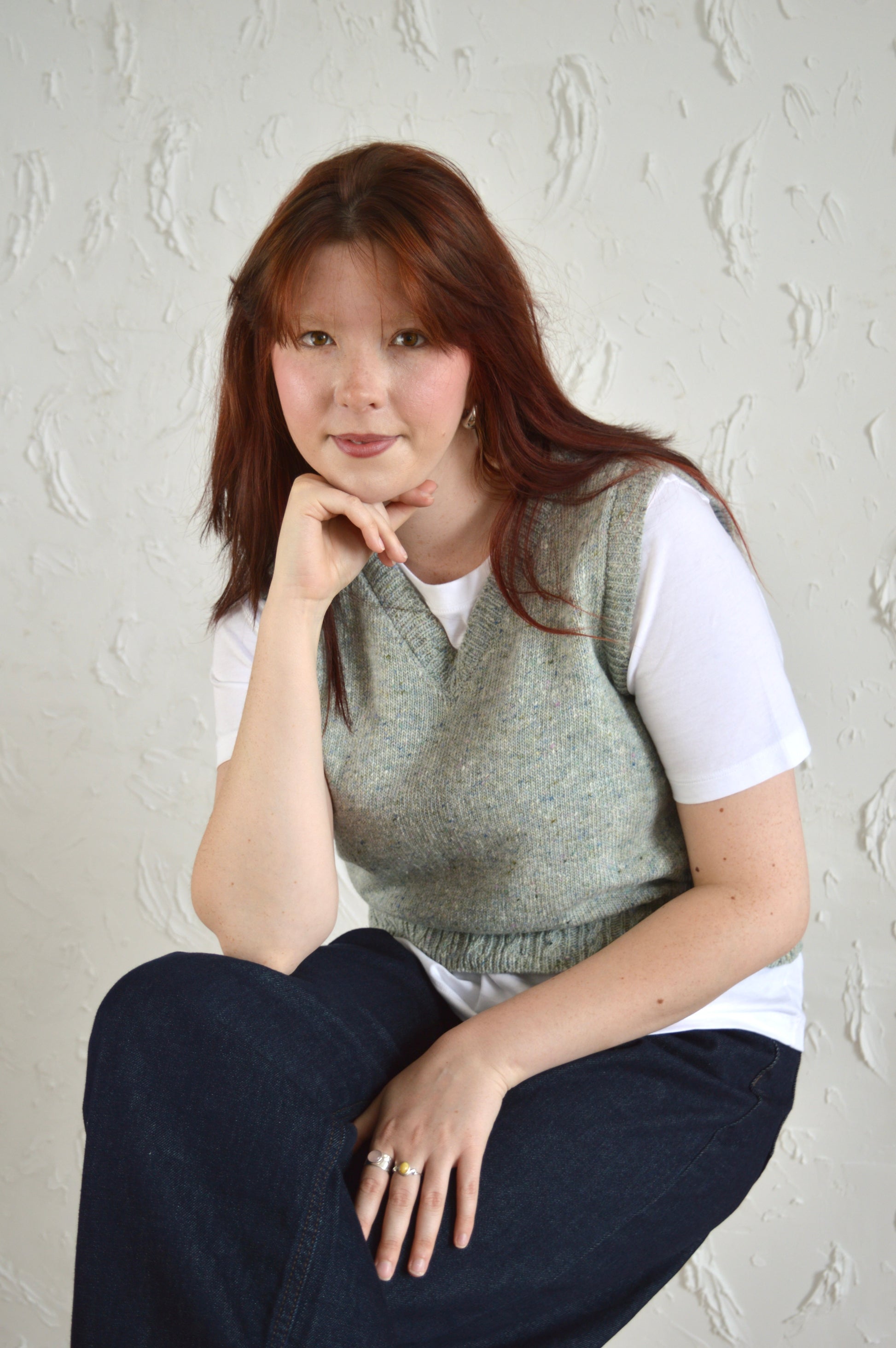 Model wearing The Cable Vest, seated on a stool with hand resting on chin in a relaxed pose.