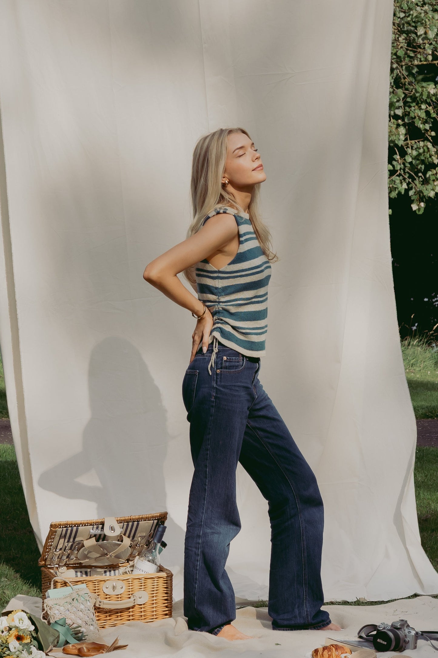 A woman stands outdoors in front of a large cream fabric backdrop, wearing a sleeveless blue and cream striped top and dark blue wide-leg jeans. A wicker picnic basket, flowers, and a camera sit on the ground nearby, with grass and trees visible in the background.