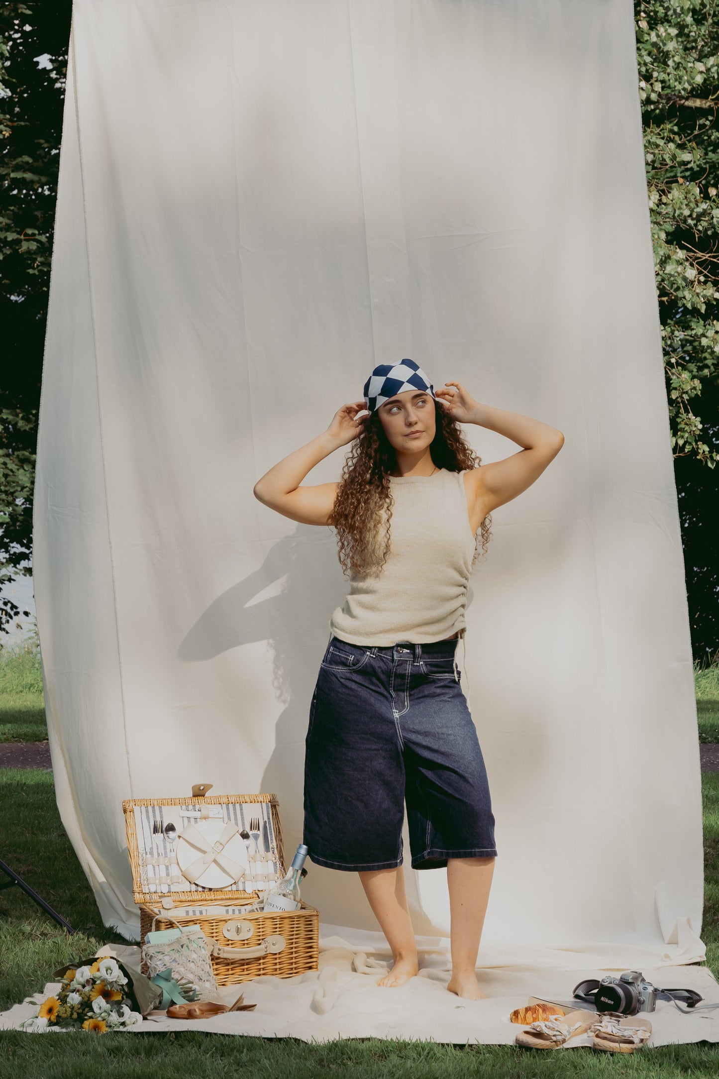 A woman stands outdoors in front of a large white fabric backdrop, wearing a beige sleeveless top, dark denim shorts, and a blue-and-white patterned headscarf. A wicker picnic basket, flowers, and shoes are arranged on the grass at her feet, with trees visible in the background.