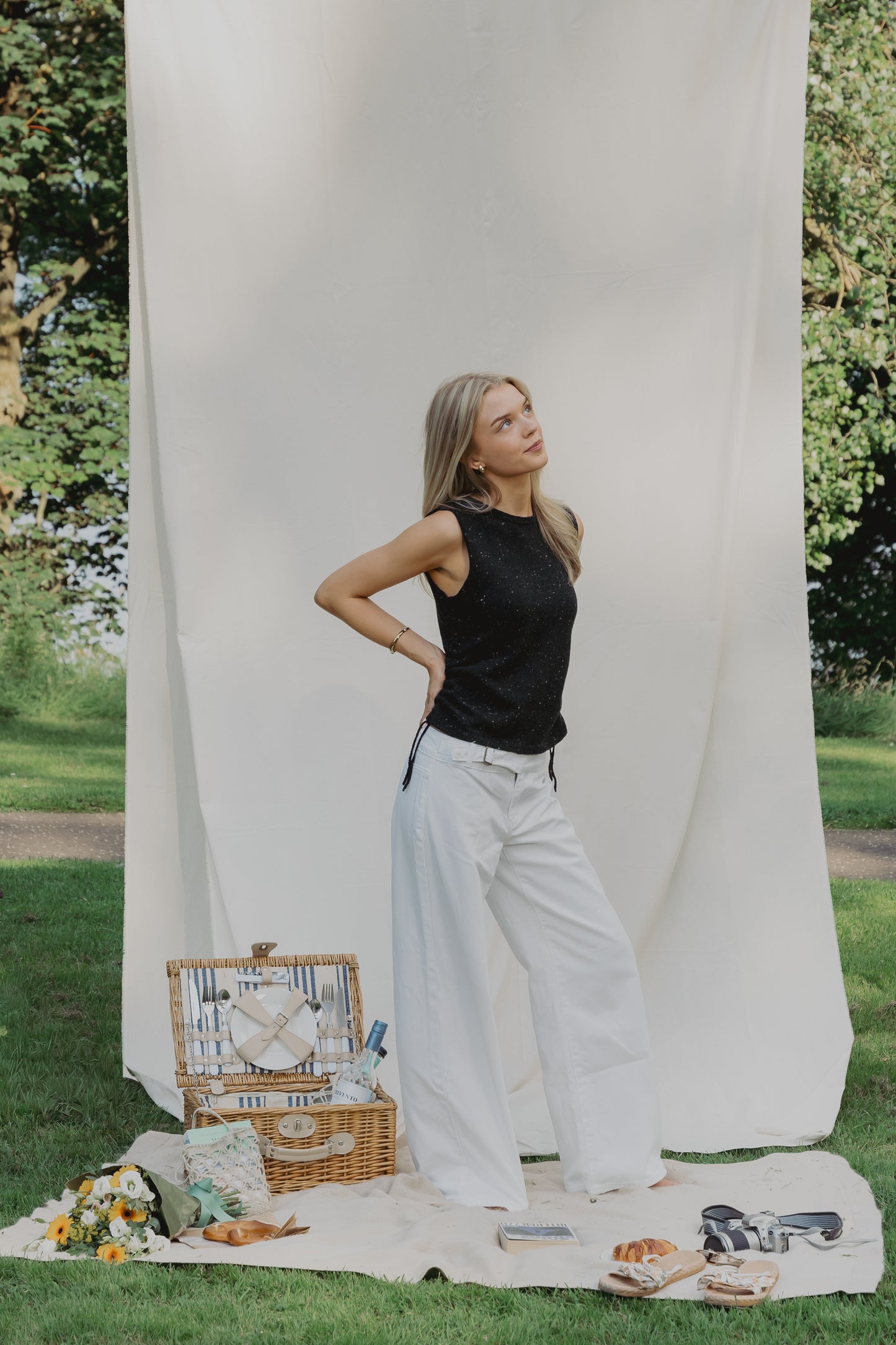 A woman stands outdoors in front of a large white fabric backdrop on grass, wearing a black sleeveless top and white wide-leg pants. A picnic basket, blanket, flowers, and sandals are arranged nearby, with trees visible in the background.
