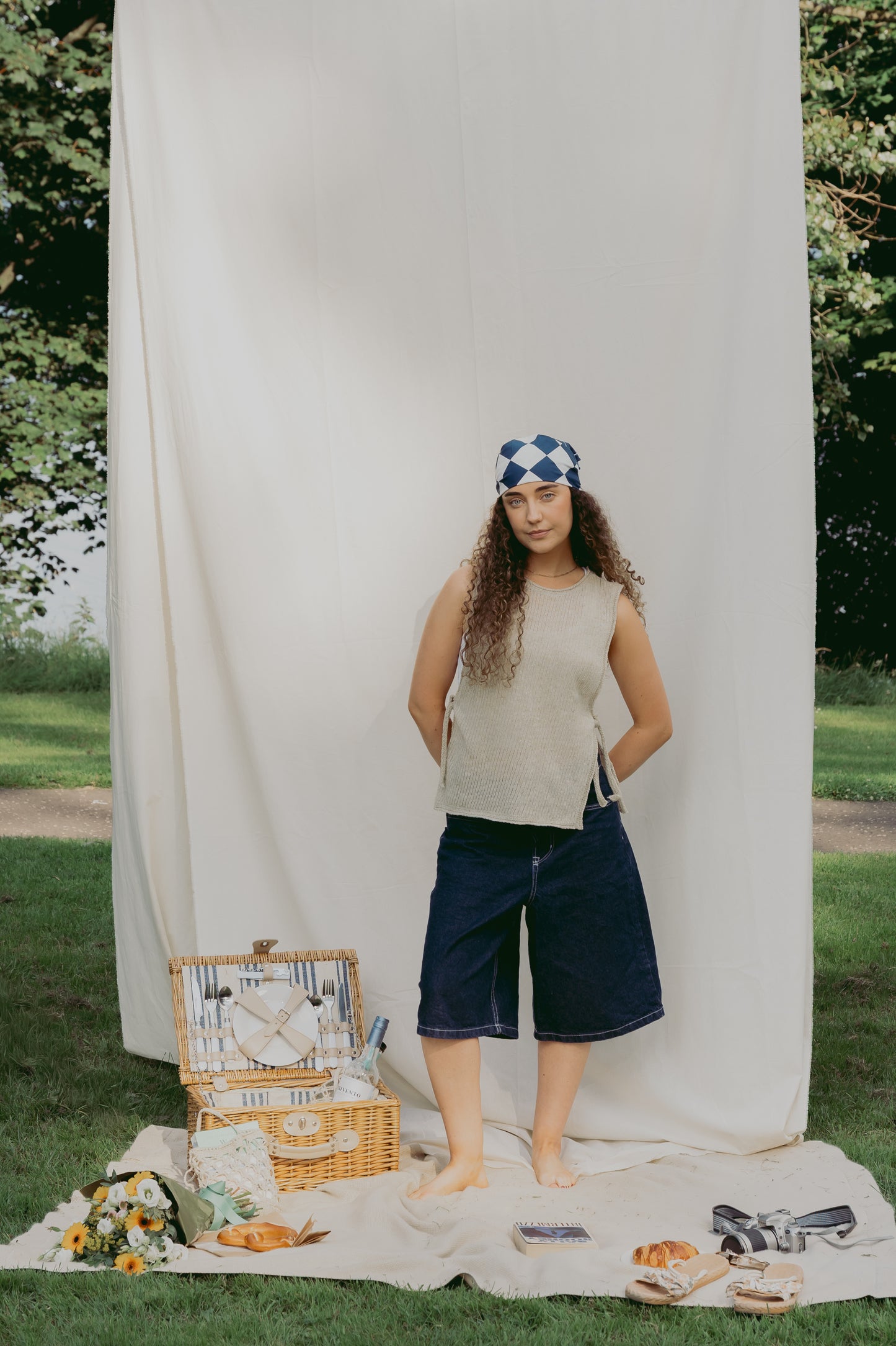 A woman stands outdoors in front of a large white fabric backdrop on grass, wearing The Basket Vest in cream, dark navy shorts, and a blue-and-white patterned headband. A picnic basket, blanket, flowers, and bread are arranged on the ground nearby.