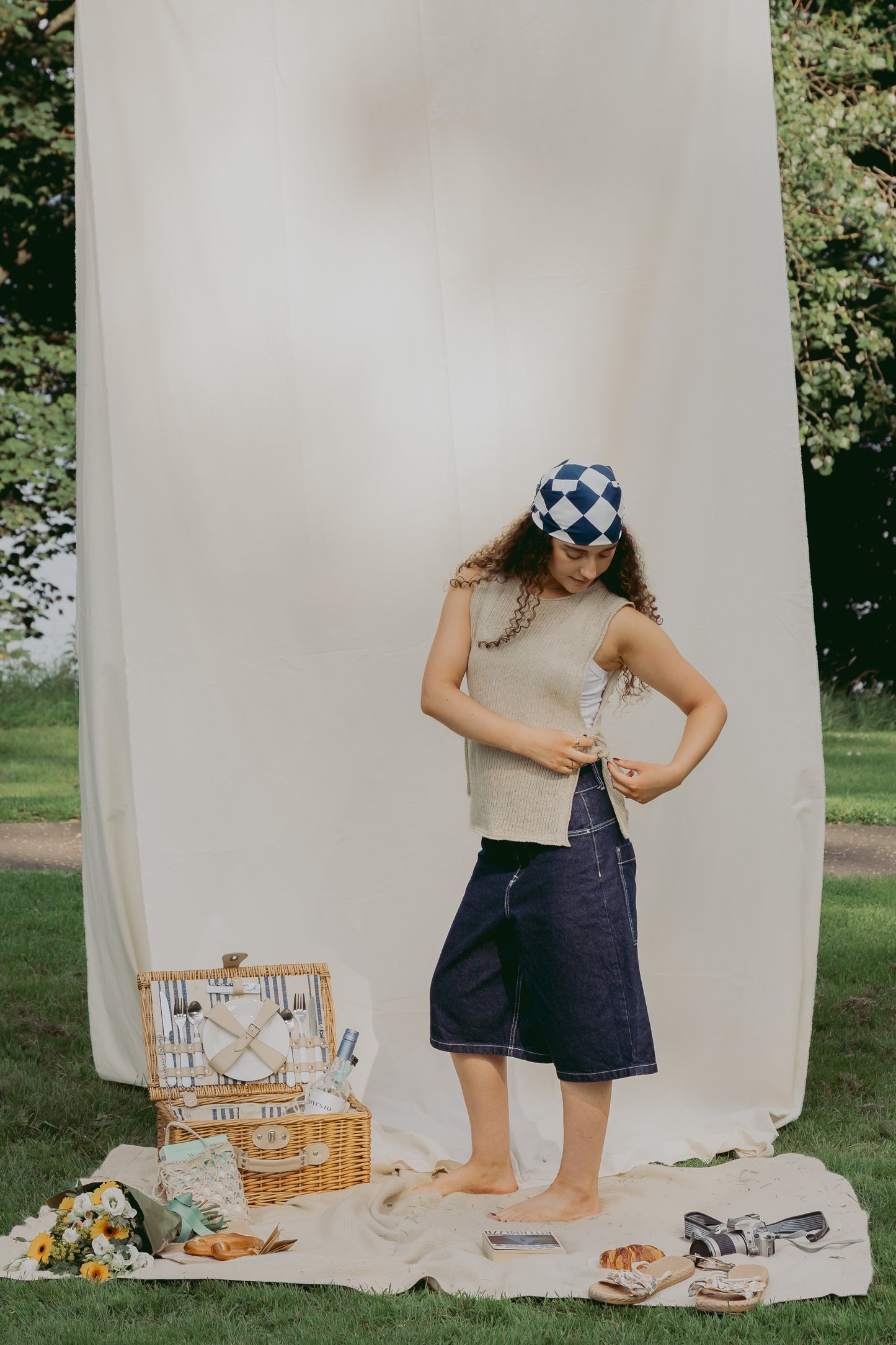 A woman stands outdoors in front of a large white fabric backdrop on grass, wearing The Basket Vest in cream, dark denim shorts, and a blue-and-white checkered headscarf. A picnic basket, flowers, and shoes are arranged on the blanket beside her.
