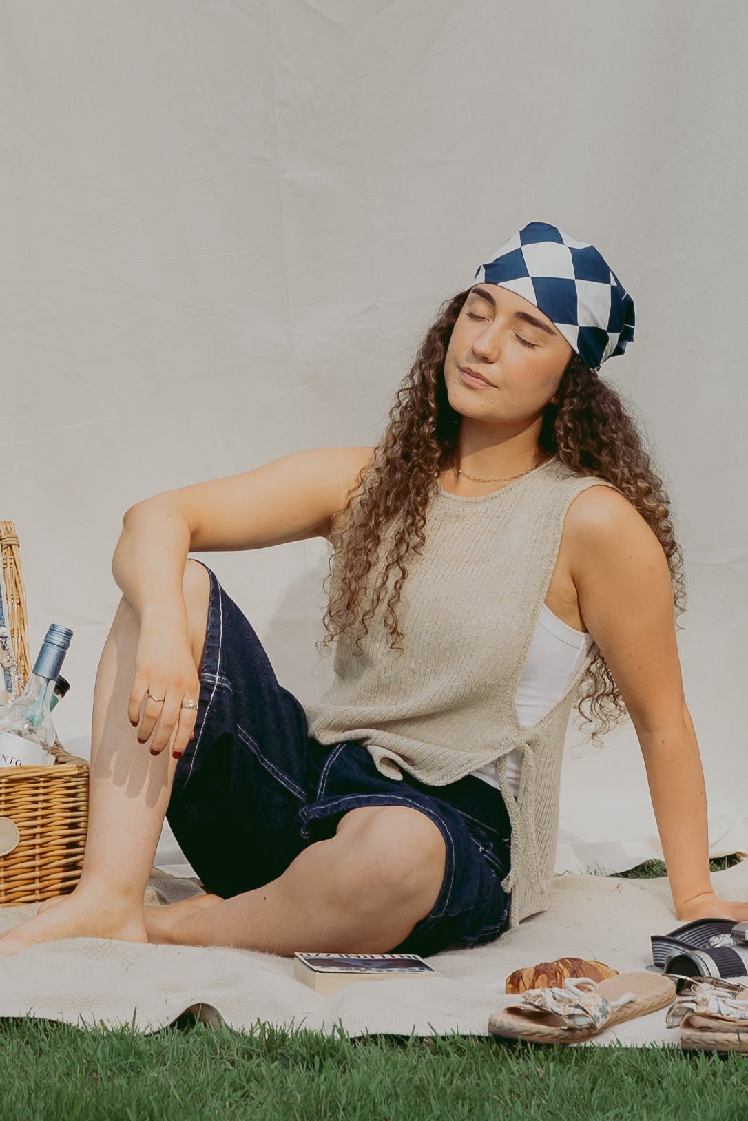 A woman sits on a cream picnic blanket outdoors on a grassy lawn, wearing The Basket Vest, dark denim shorts, and a blue-and-white checkered headband. A wicker picnic basket, flowers, sunglasses, and sandals are arranged beside her in front of a draped white fabric backdrop.