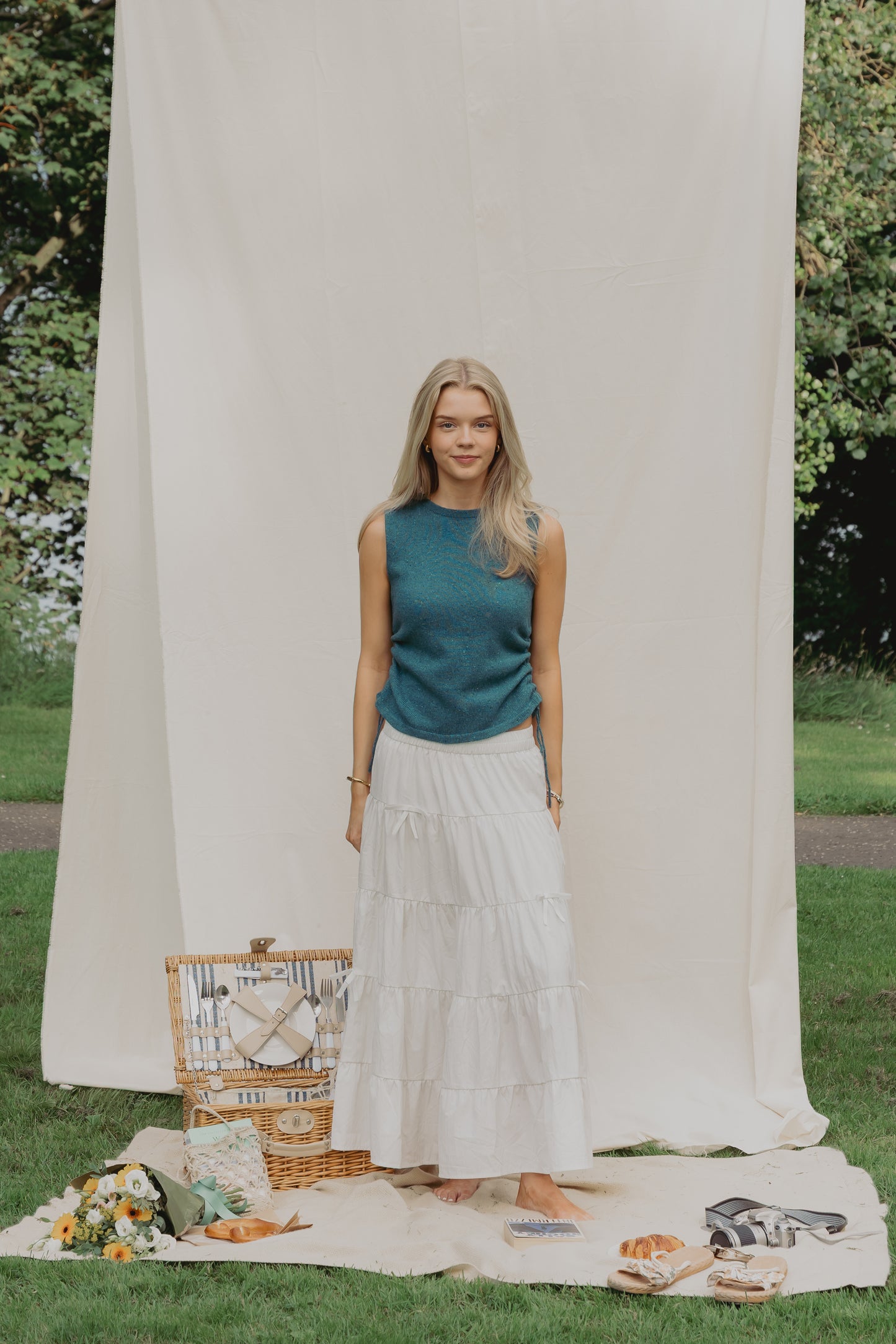 A woman stands outdoors on a blanket in front of a large cream fabric backdrop, wearing a sleeveless teal top and a long white tiered skirt. A picnic basket, flowers, bread, and sandals are arranged on the grass beside her.