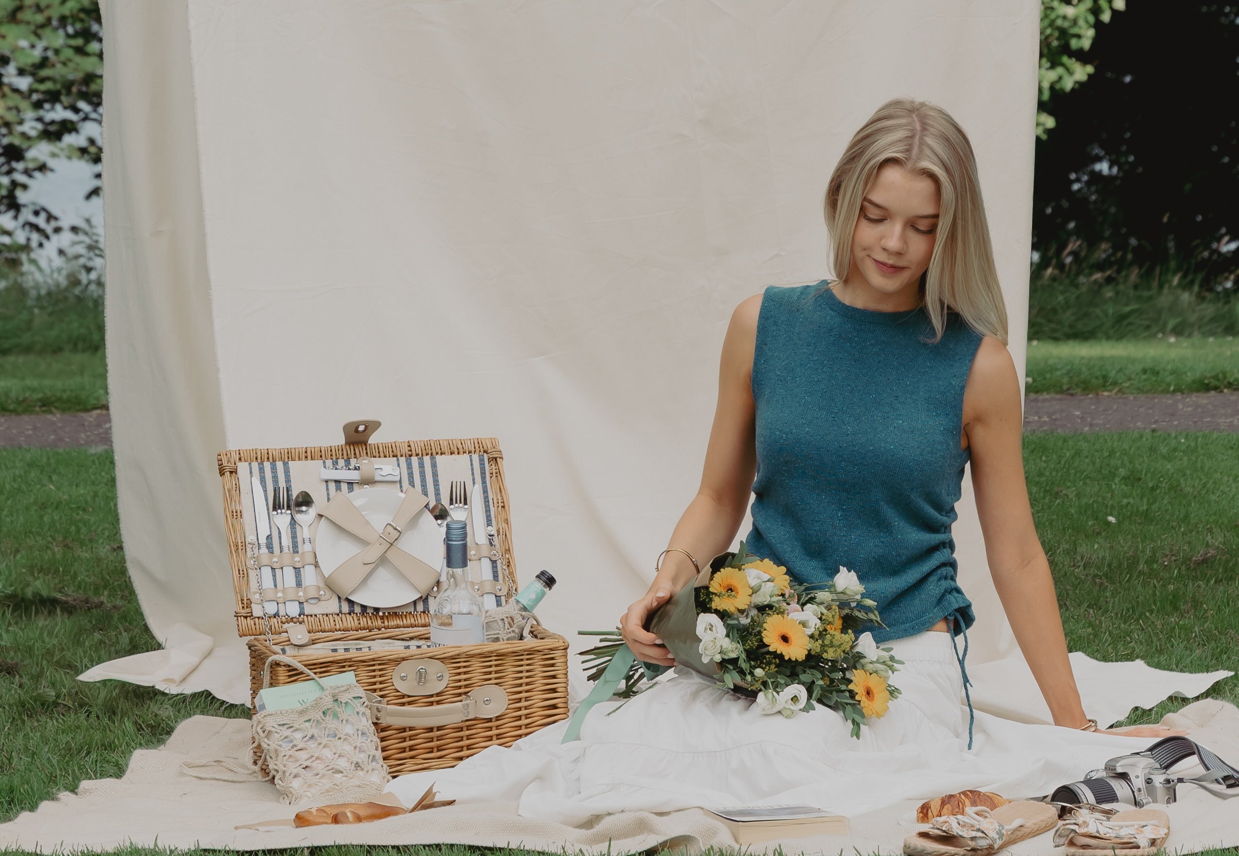 A woman sits on a cream picnic blanket on a grassy lawn, holding a bouquet of yellow and white flowers. To her left is an open wicker picnic basket with plates, bottles, and bread, and her white dress is spread out beneath her. She is wearing The Blacket Top in teal