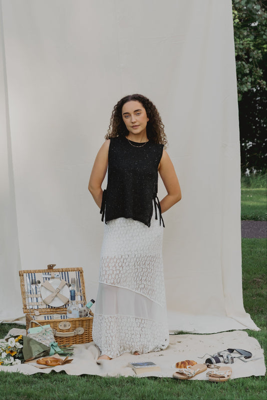 A woman stands outdoors in front of a large white fabric backdrop on grass, wearing The Basket Vest in black and long white lace skirt A picnic basket, flowers, and shoes are arranged on the blanket beside her.