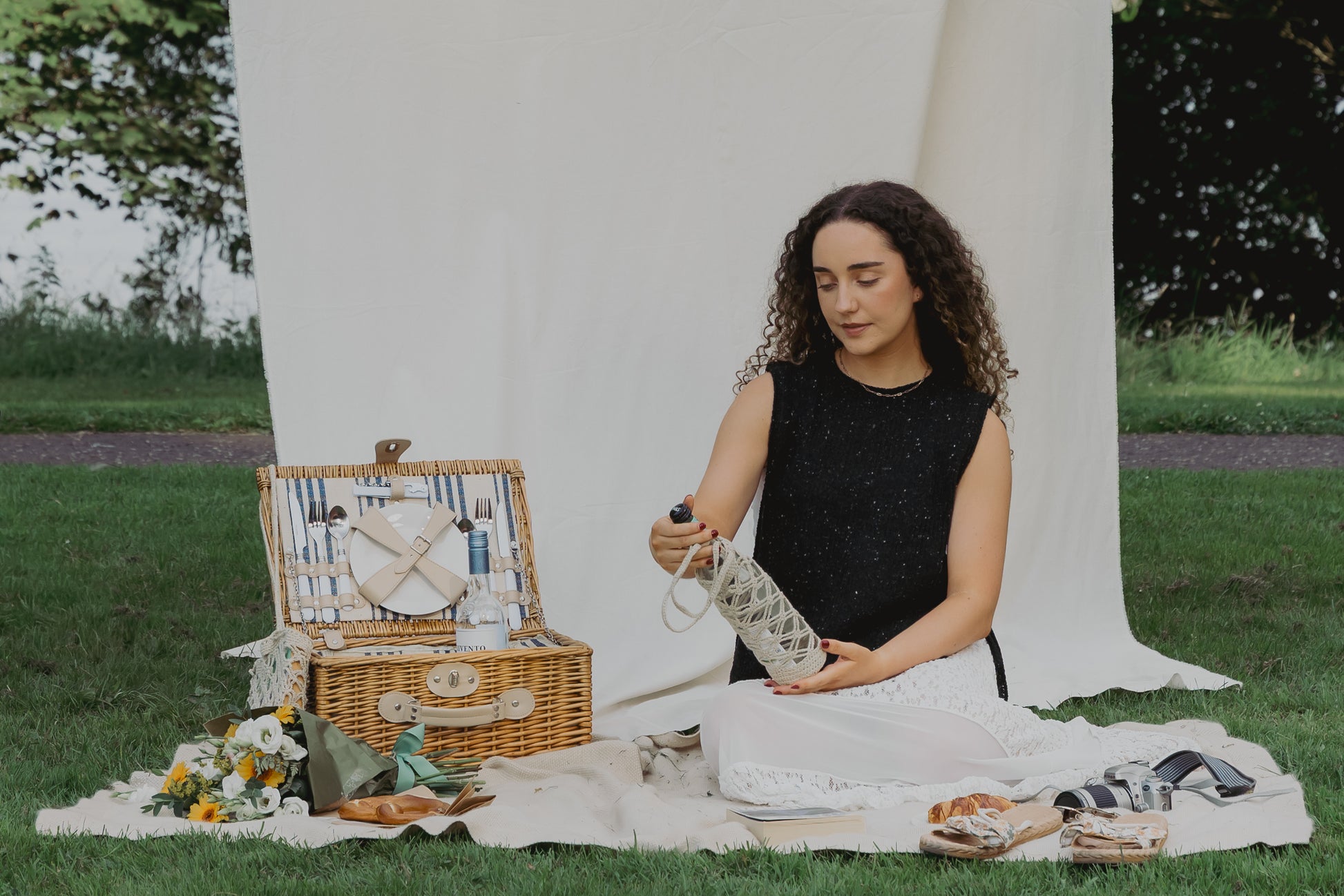 A woman sits on a white picnic blanket outdoors on a grassy lawn, holding a wine glass in The Bottle Sling. A wicker picnic basket with plates and utensils, sandals, and small accessories are arranged beside her, with a white draped backdrop behind her.