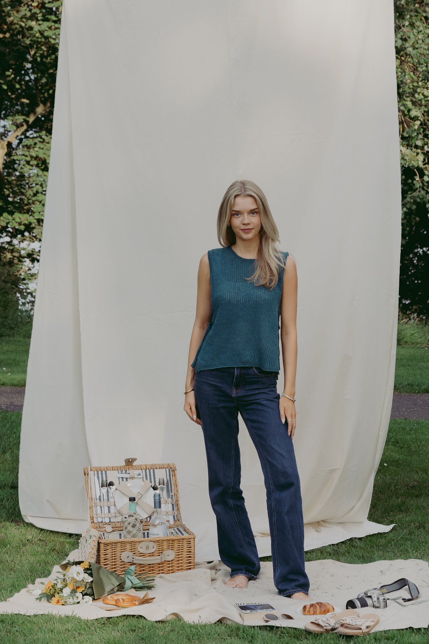 A woman stands outdoors on a picnic blanket in front of a large cream fabric backdrop, wearing The Basket Vest in teal and dark blue jeans. A wicker picnic basket with plates and bread is on the grass beside her.