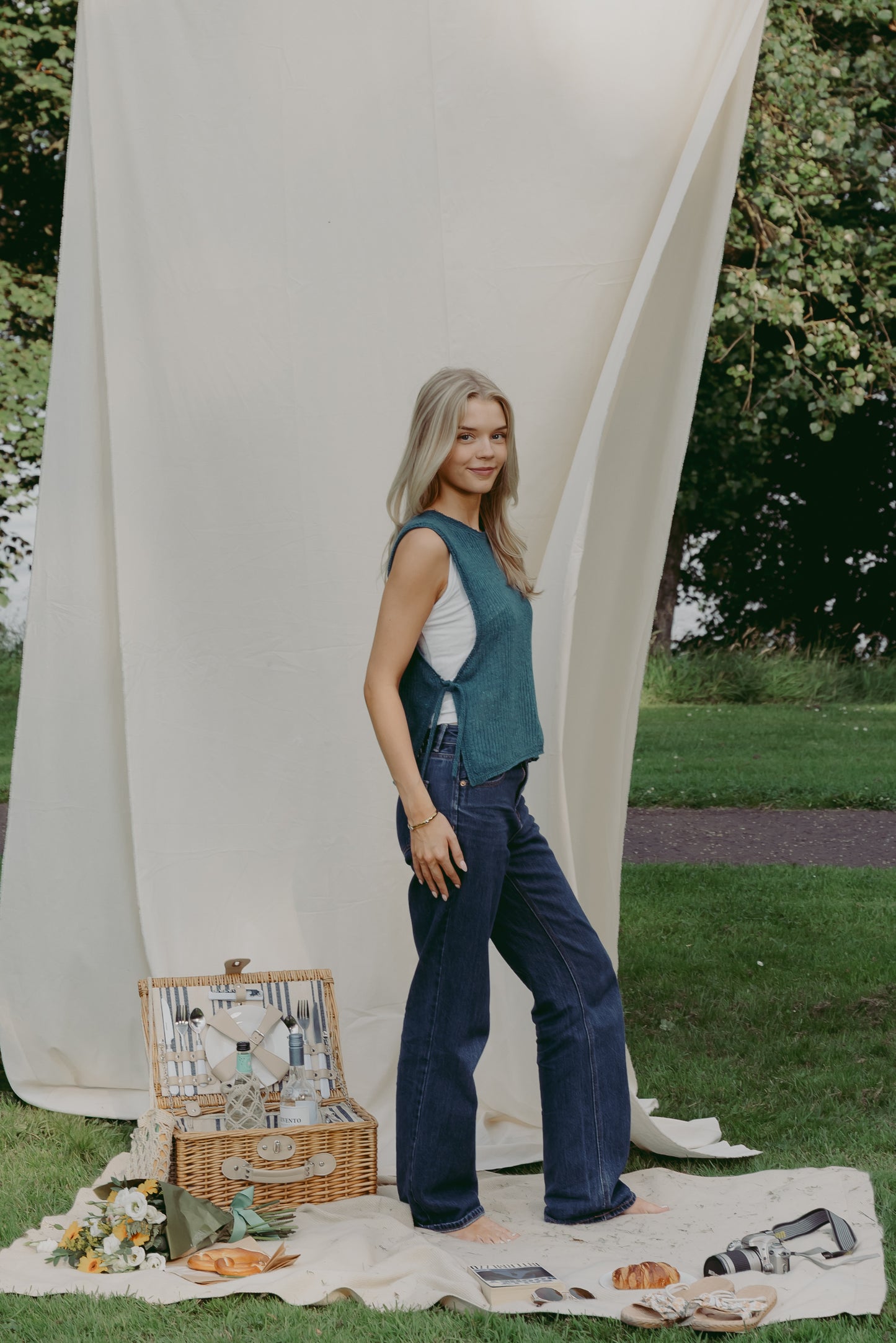 A woman stands outdoors on a cream picnic blanket with a wicker picnic basket, wearing The Basket Vest in teal, over a white top and dark blue wide-leg jeans. A large cream fabric backdrop hangs behind her in a park setting with grass and trees.
