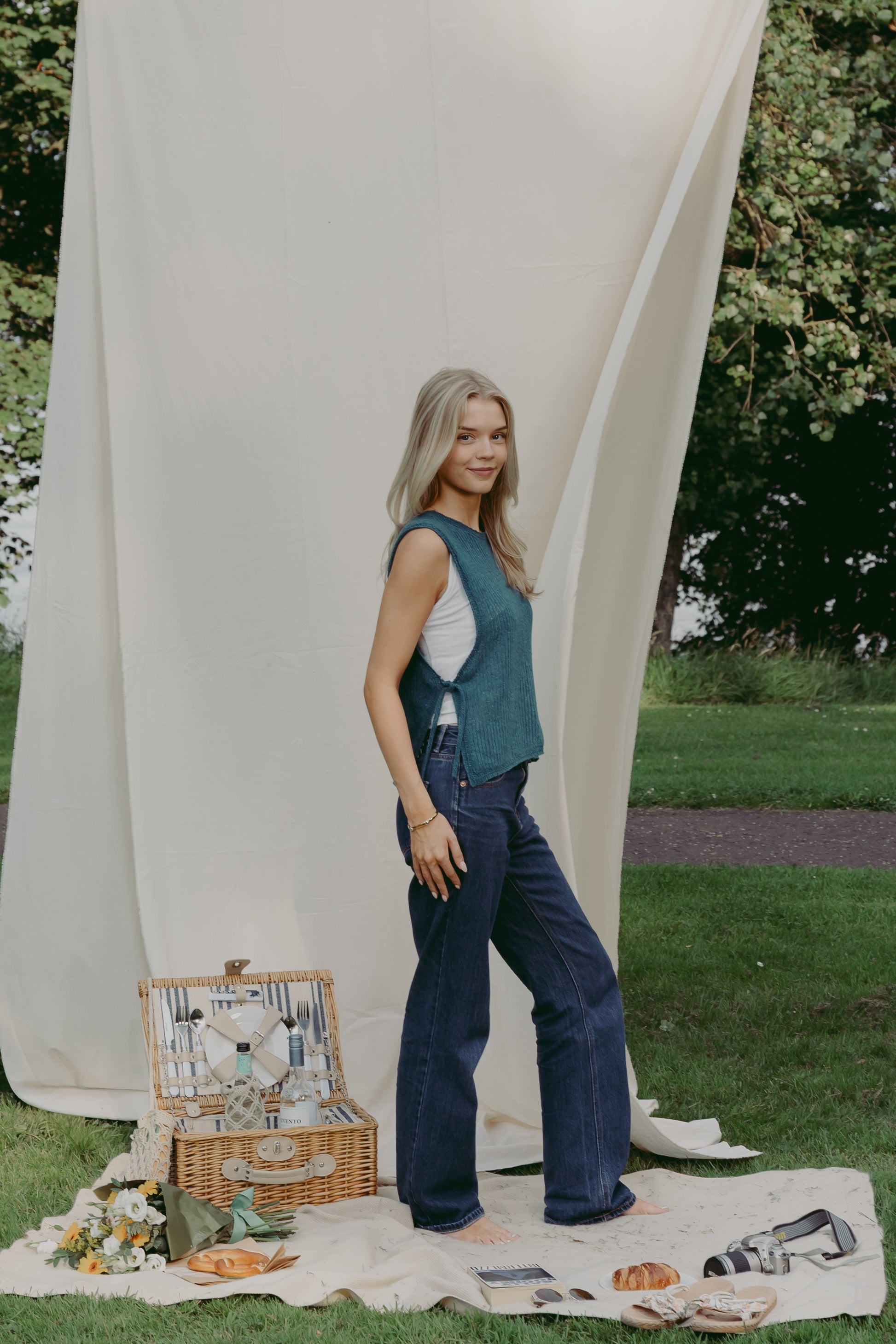 A woman stands outdoors on a cream picnic blanket with a wicker picnic basket, wearing The Basket Vest in teal, over a white top and dark blue wide-leg jeans. A large cream fabric backdrop hangs behind her in a park setting with grass and trees.