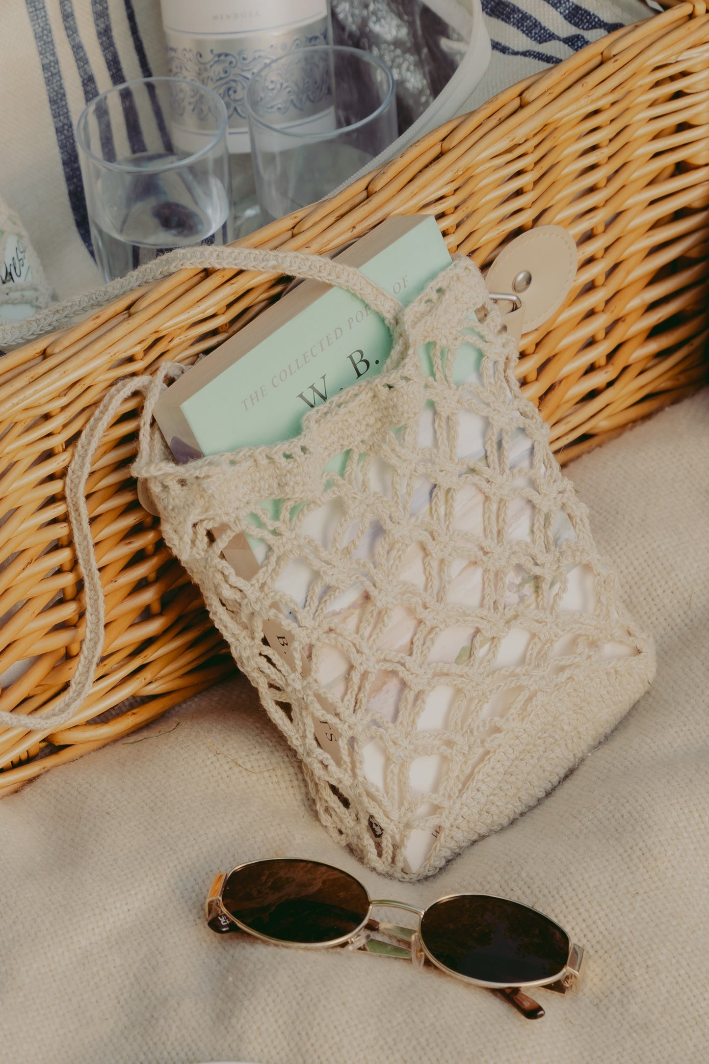 A cream-colored crocheted drawstring bag with a handle, styled on a beige fabric surface next to a wicker tray, sunglasses, and a book.