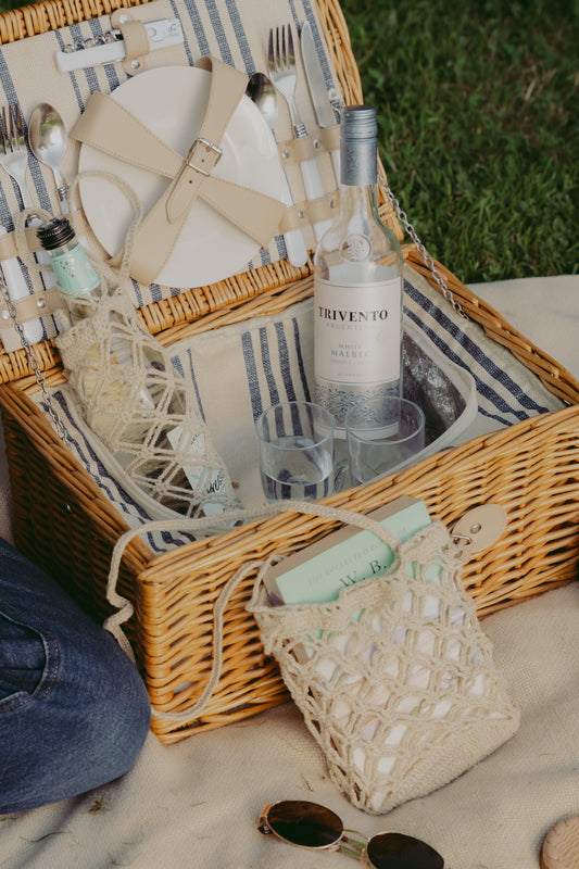 A wicker picnic basket filled with plates, cutlery, napkins, and a bottle in The Bottle Sling, styled outdoors on a blanket with sunglasses, a crocheted drawstring bag, and part of a person's jeans visible.