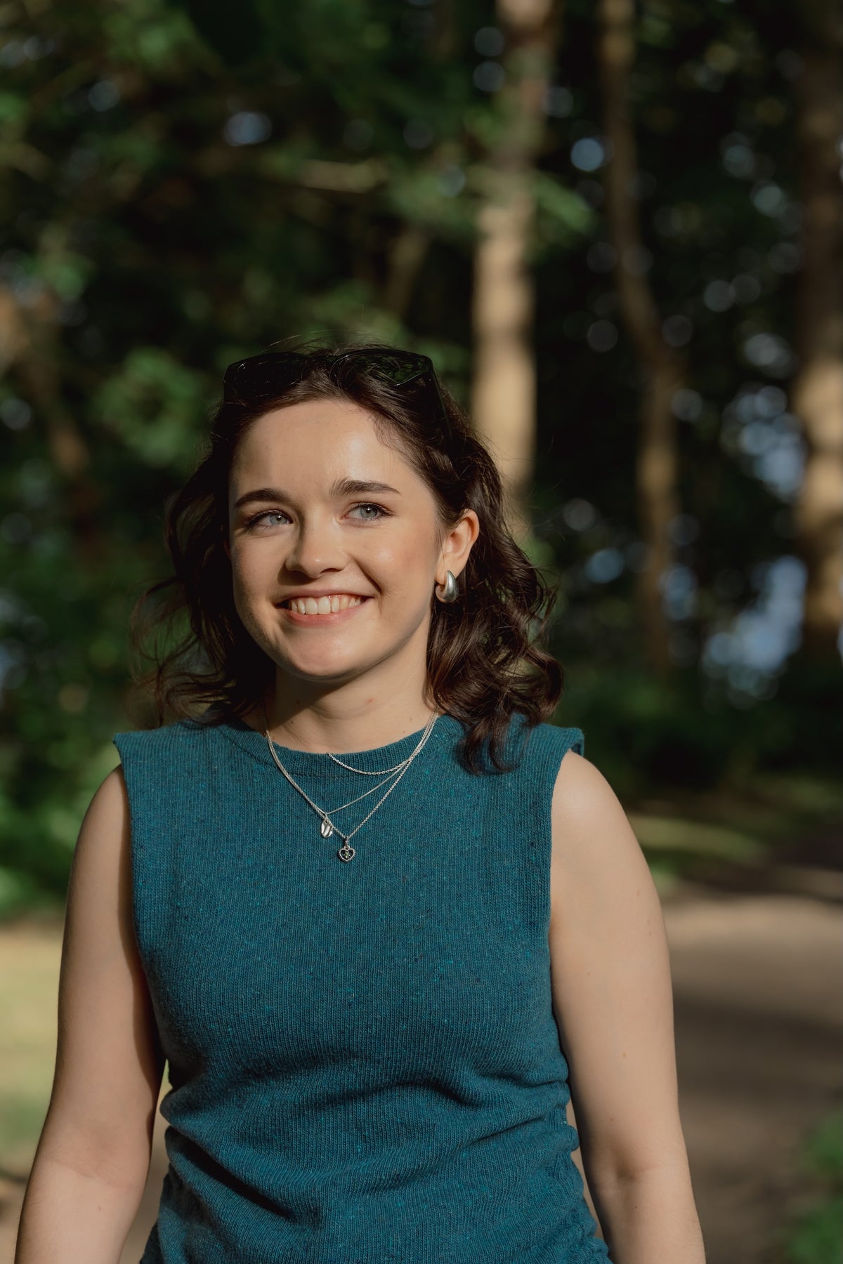 A smiling Caitlin outdoors wearing The Blanket Top in teal and layered silver necklaces, with trees and a path blurred in the background.