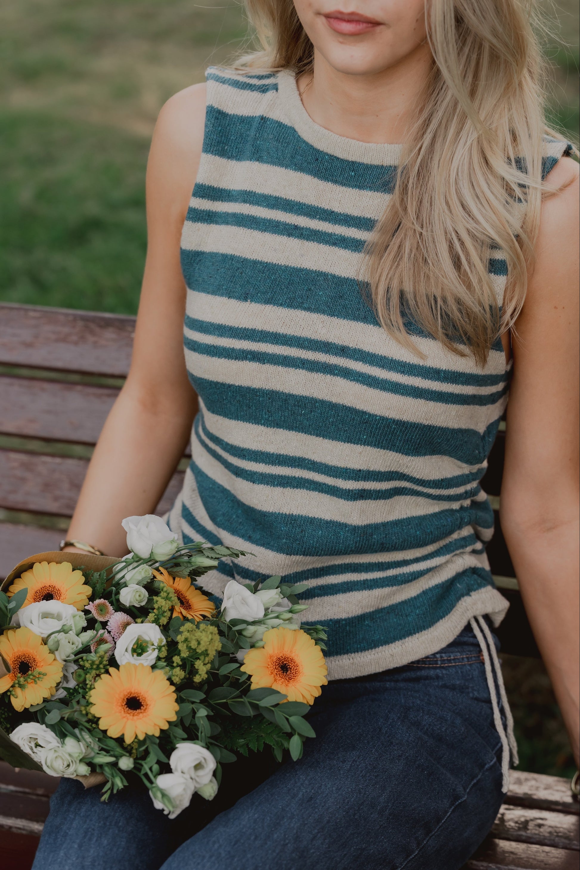 A woman sitting on a wooden bench holding a bouquet of yellow and white flowers, wearing The Blanket Top with horizontal teal and cream stripes and dark blue jeans.